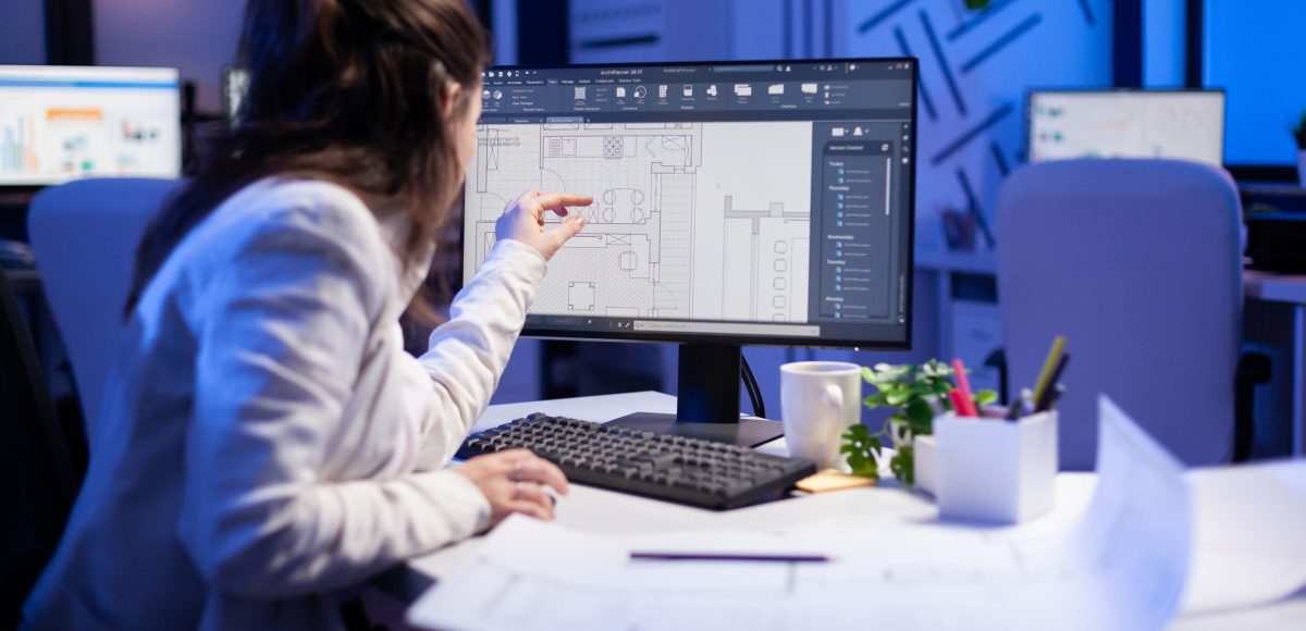 Over the shoulder shot of engineer woman drawing architectural plans and looking at cad software on desktop computer. Designer using arhitecture blueprints of buildings creating industrial prototype