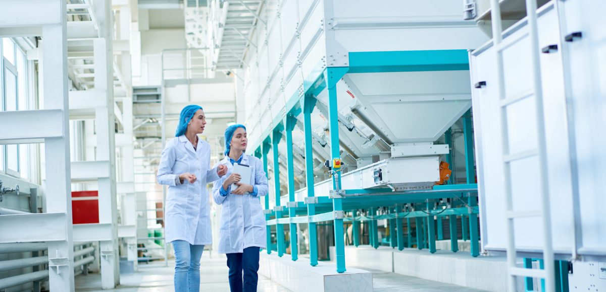 Full length portrait of  two young female workers wearing lab coats walking by  power units crossing hall in  in clean production workshop, copy space
